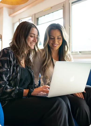 Two women sitting together smiling and looking at a laptop screen.