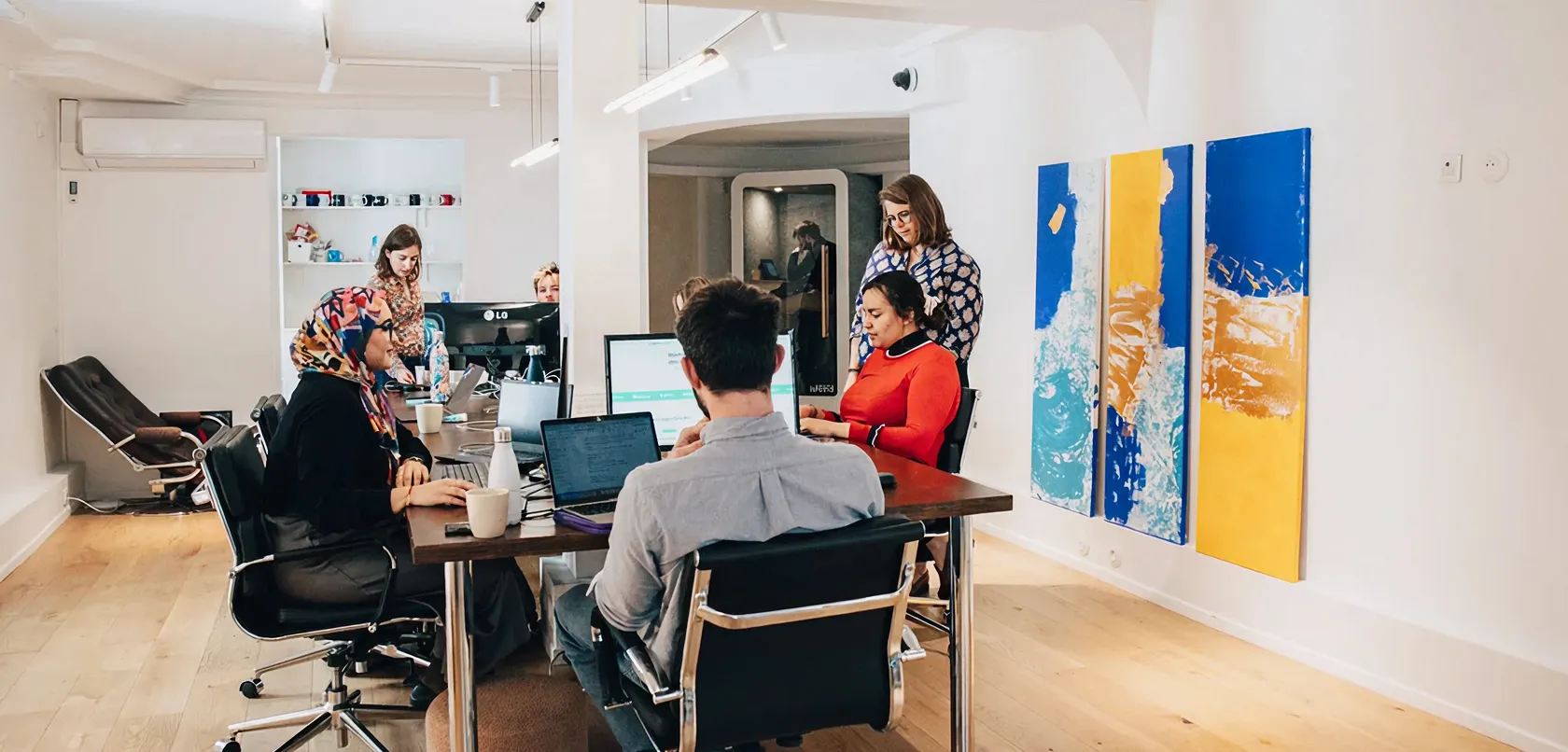 Group of diverse colleagues working and collaborating around a table in a modern office with abstract blue and yellow artwork on the wall.