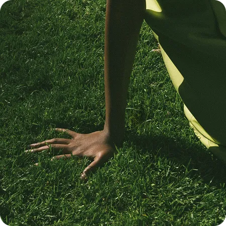Close-up of a dark-skinned hand with rings resting on green grass, with part of a person in light green clothing visible.
