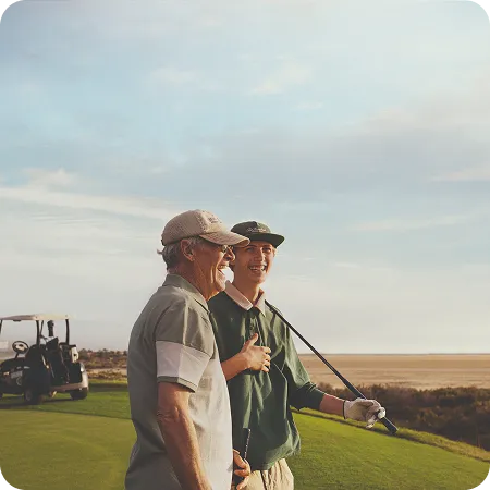Two men standing on a golf course, smiling and holding golf clubs, with a golf cart in the background.
