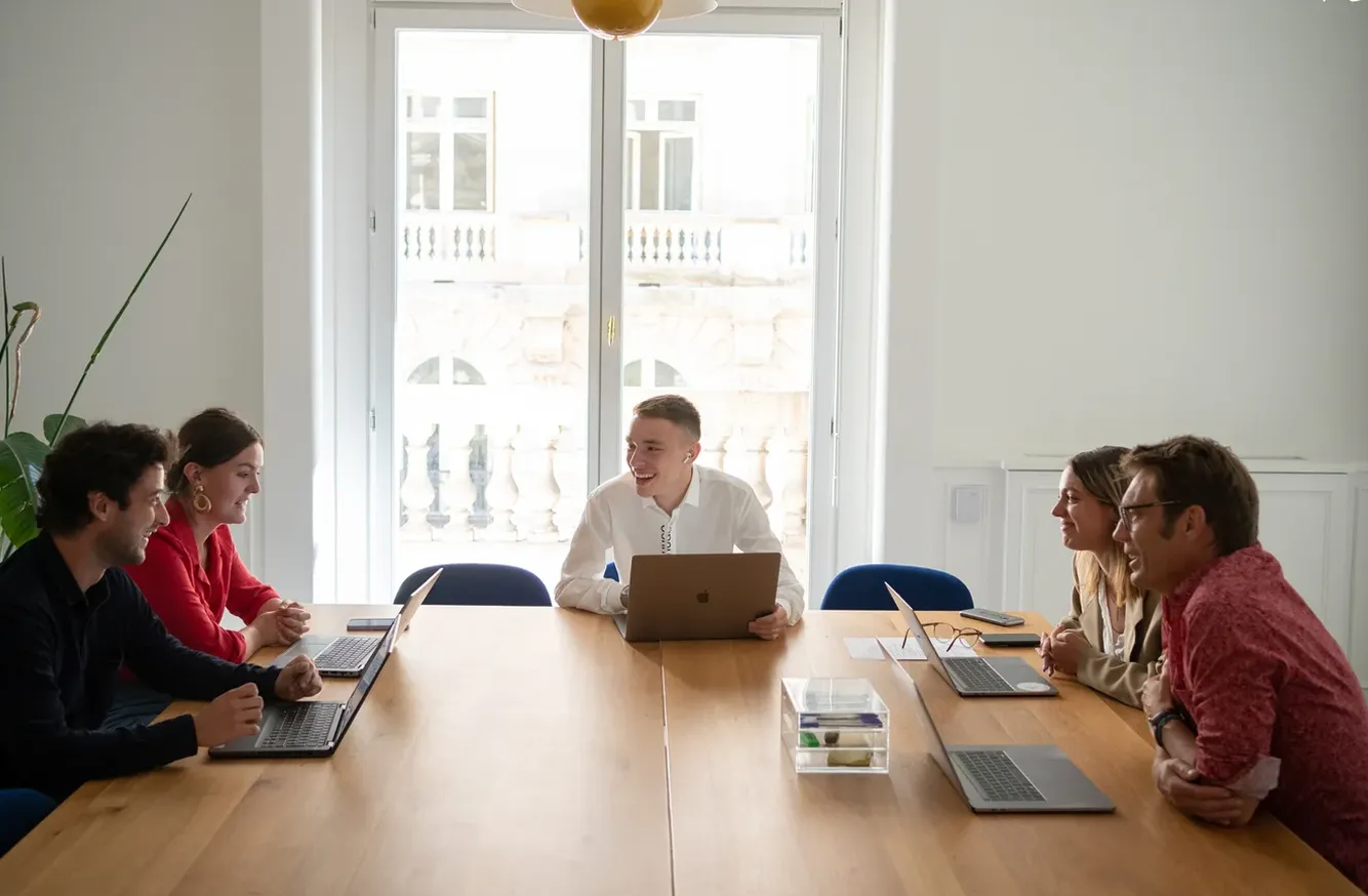 Five people sitting around a wooden table in a bright meeting room, smiling and chatting with laptops open.