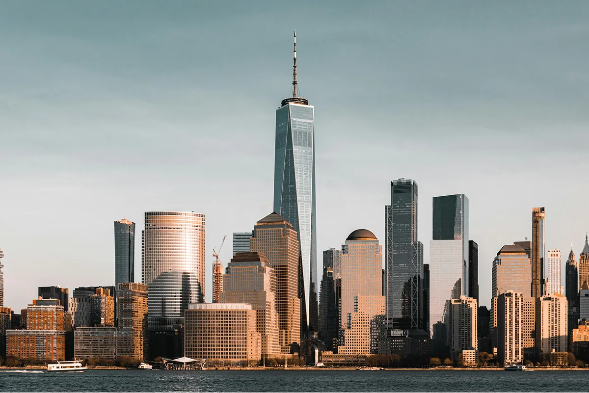 Manhattan skyline with One World Trade Center towering above other buildings, viewed across the water at sunset.