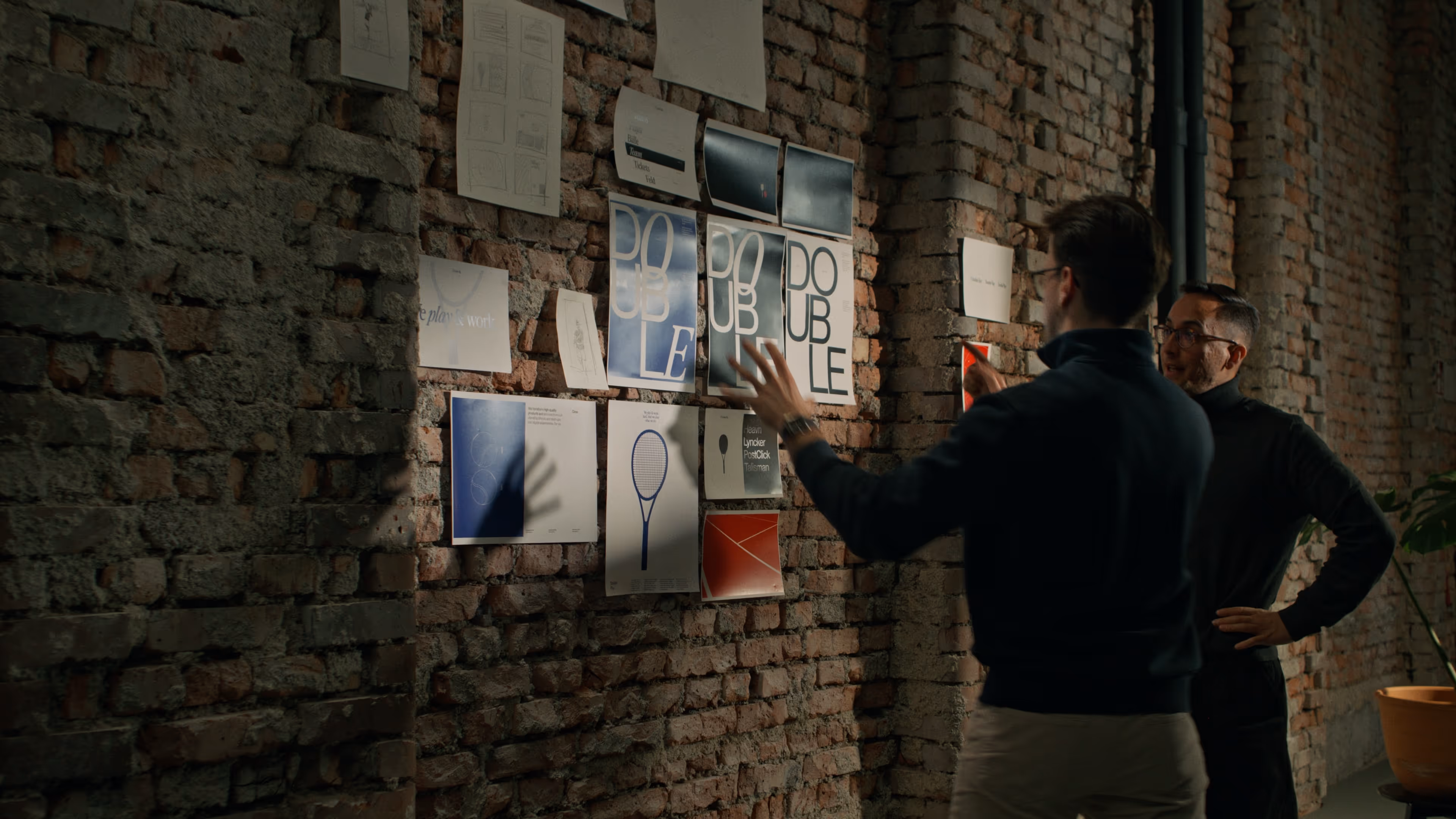 Two man standing in front of a brick wall full of paper posters in a discussion