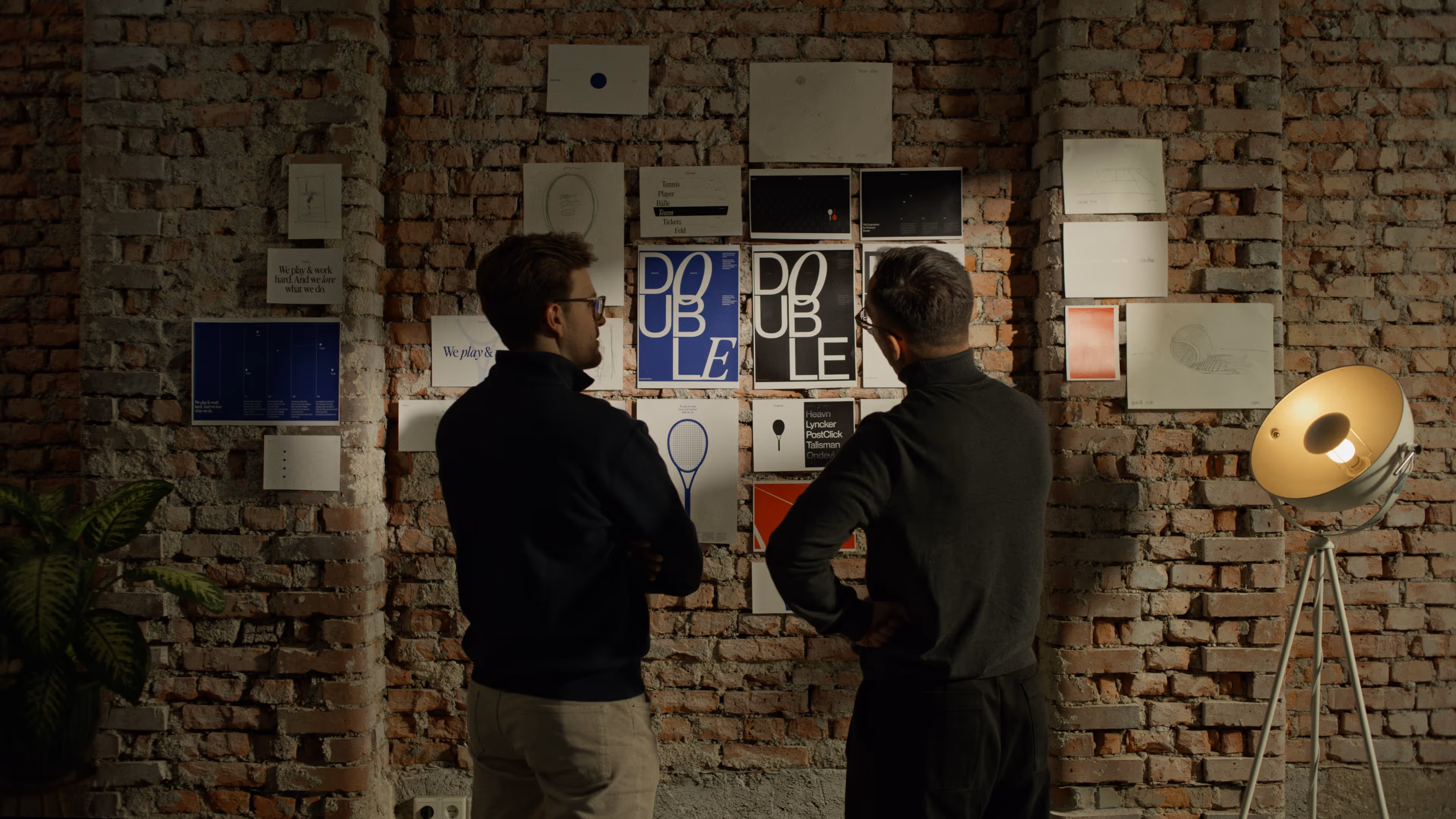 Two man standing in front of a brick wall full of paper posters in a discussion