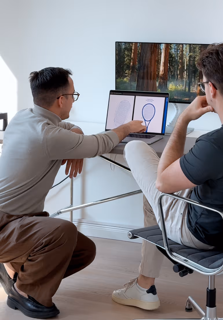 Two men discuss a laptop screen showing tennis racket designs in a modern office setting.