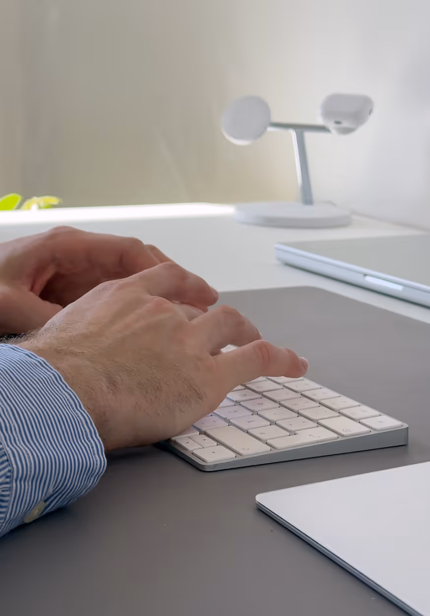 Hands typing on a slim white keyboard on a desk with a laptop and a small white lamp in background.