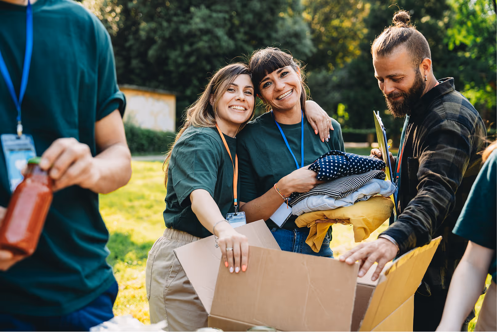 Two smiling women with volunteer badges packing clothes into a box outdoors, with a man handling the box nearby.