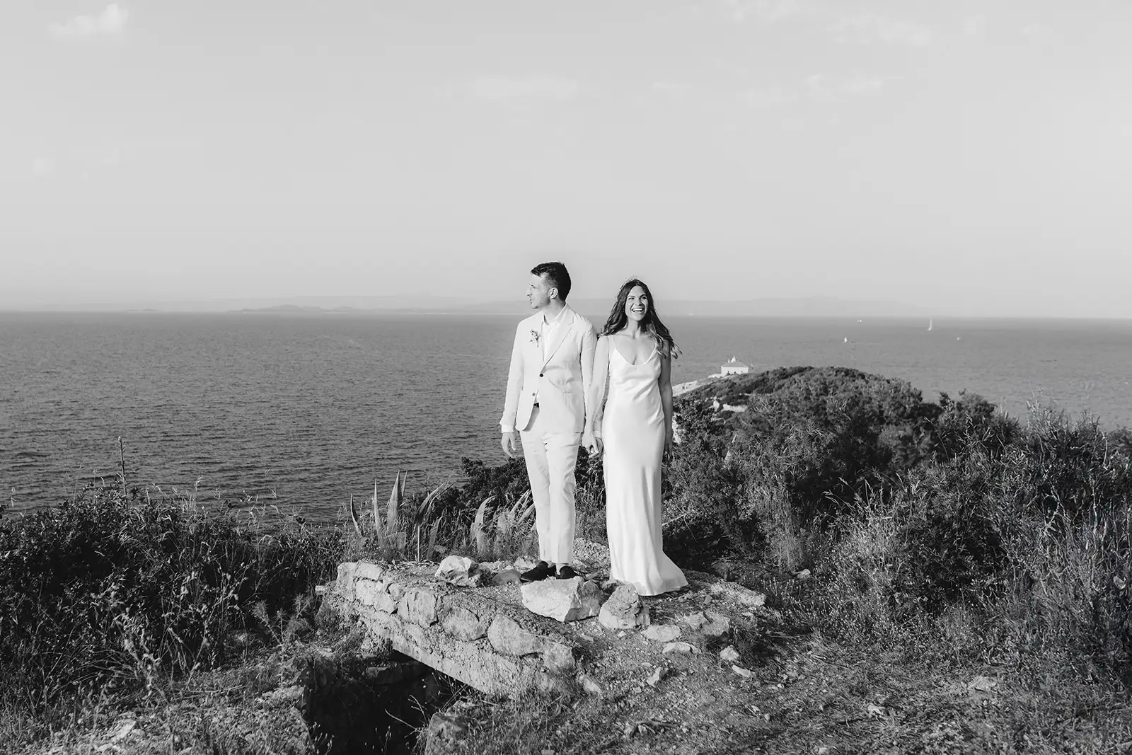 Bride and groom on a limestone wall at the cliff edge of Fort George, Vis, the lighthouse islet across the Adriatic behind them. A Fort George wedding portrait in black and white.