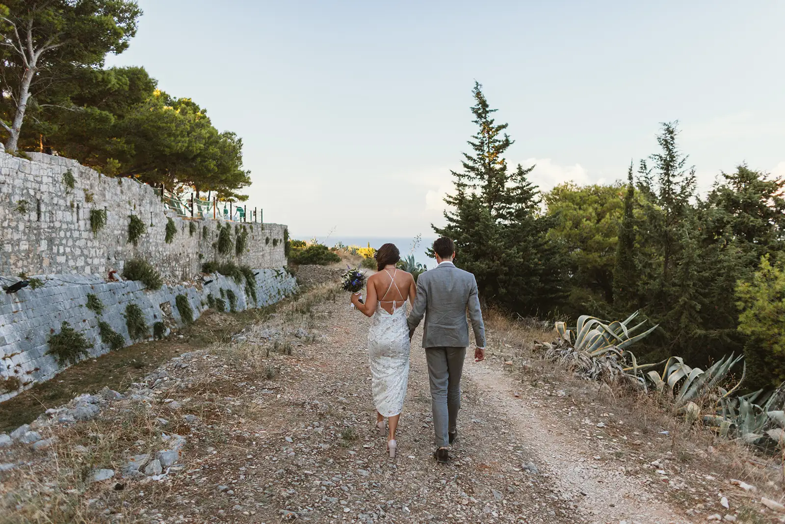 Bride and groom walk a hillside path at the Fort George wedding venue above the Adriatic on Vis, sea view ahead.