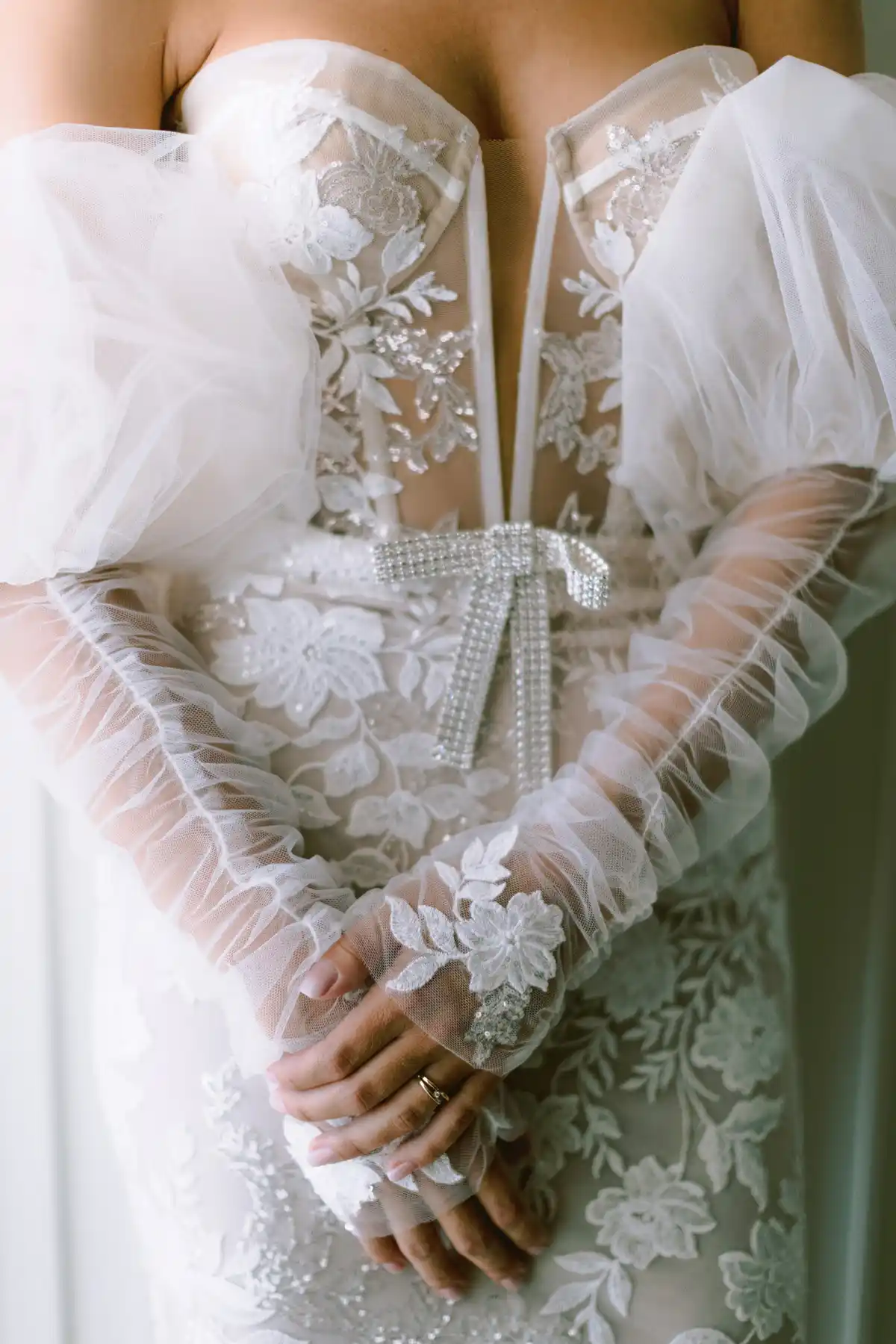 Clasped hands, sheer tulle sleeve, floral lace appliqué, crystal bow at the waist, ring visible