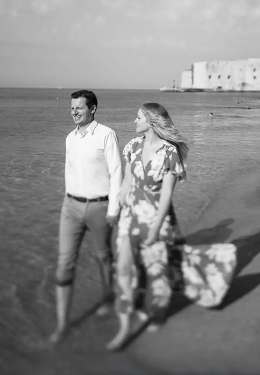 Black and white, Megan and Alex walking hand-in-hand by the sea, her hair windblown, Dubrovnik Old Town beyond