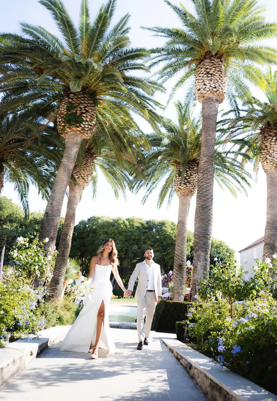 Andrea and Marko walking hand in hand along a sunlit palm-lined path, pool behind them, Hotel Lemongarden, Brač