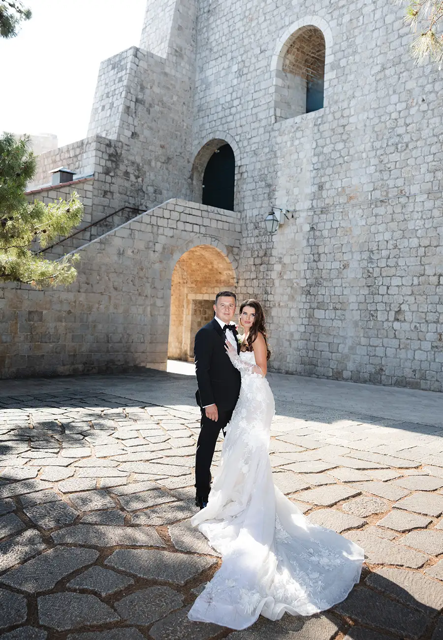 Zekira and Emin standing against a stone fortress wall, arched openings, external staircase, cobblestones underfoot, Dubrovnik
