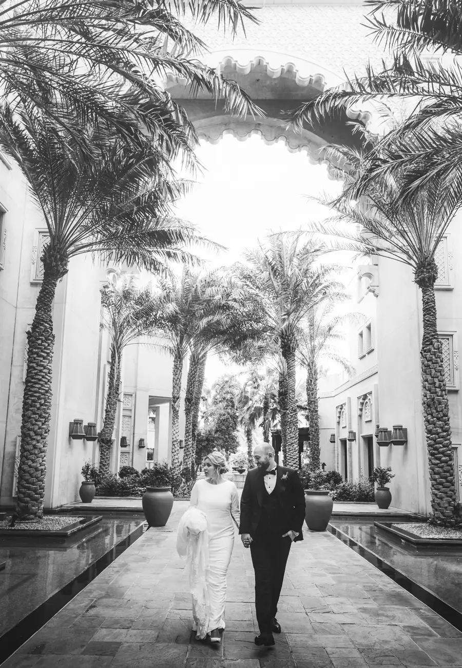 Black and white, Mariam and Ivan walking hand-in-hand through a symmetrical palm-lined courtyard with reflecting pools, Dubai