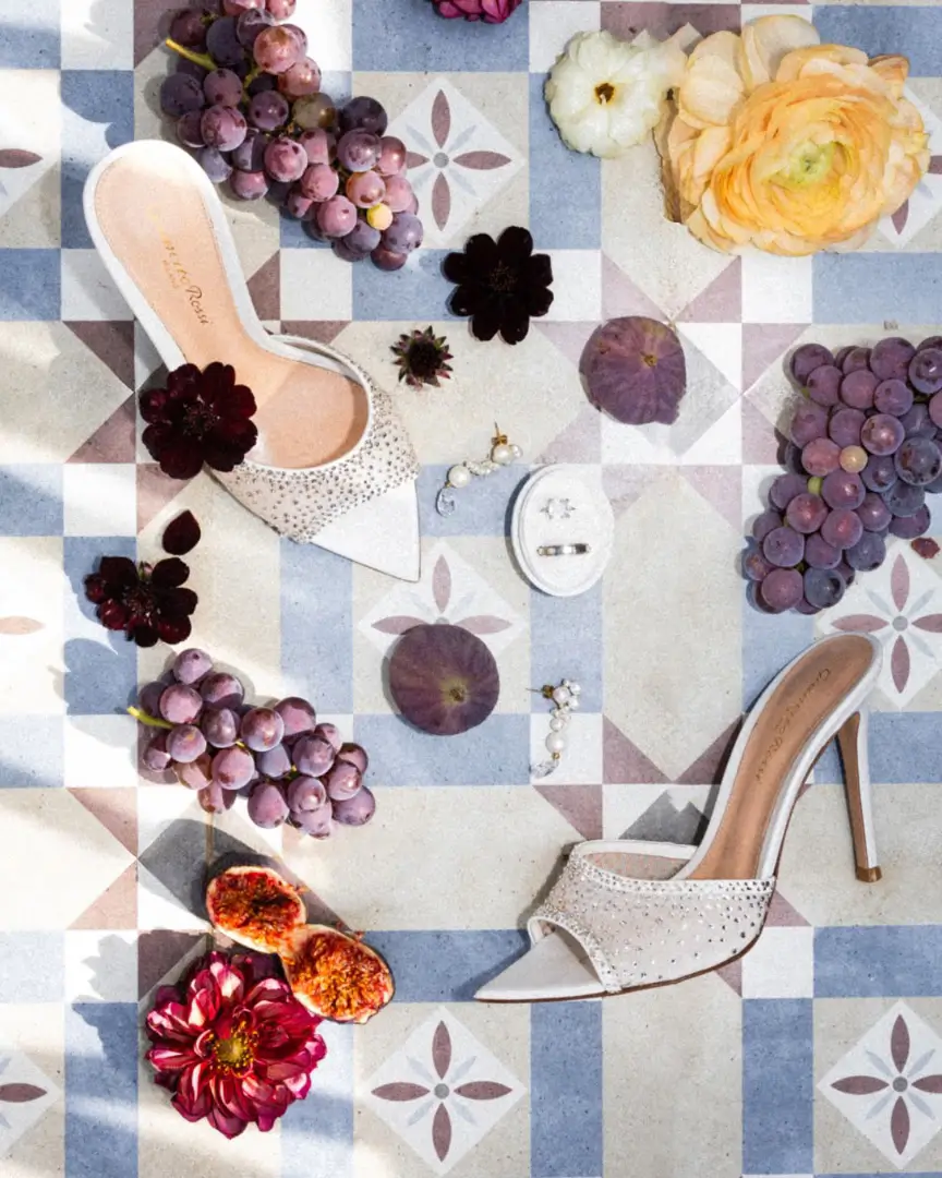 White heeled sandals, pearl earrings, a ring box, burgundy flowers, figs, and grapes on mosaic tile