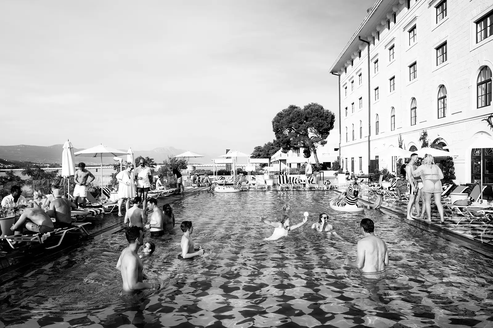 Black and white, guests swimming in Brown Beach House's iconic pool, bride in white among them, columned building behind, Trogir