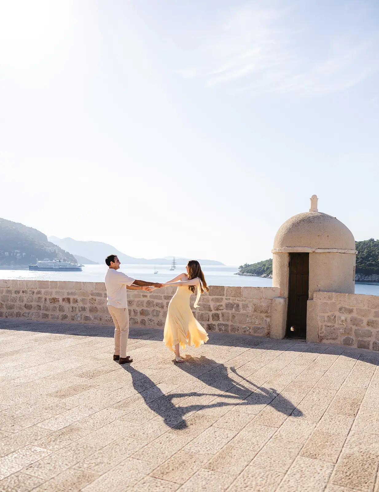 A man turns a woman on a stone terrace above the sea, a domed turret beside them, Dubrovnik.