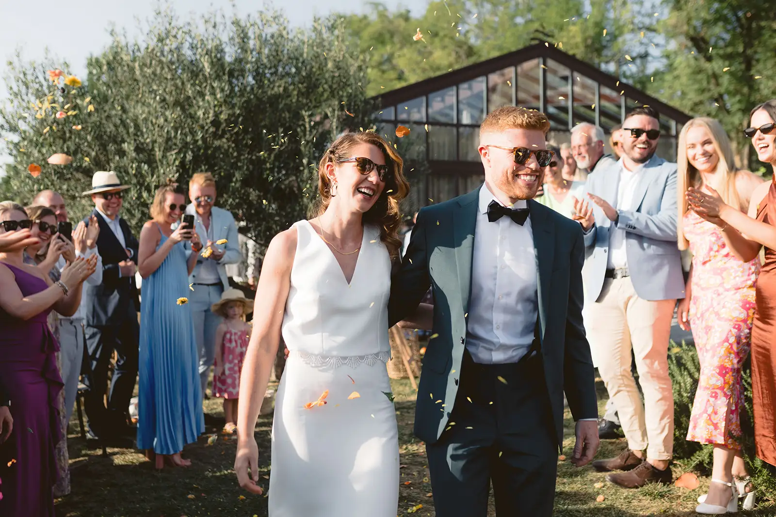 A couple walk through a corridor of guests throwing orange-and-yellow petals, a glasshouse behind them, San Canzian, Istria.