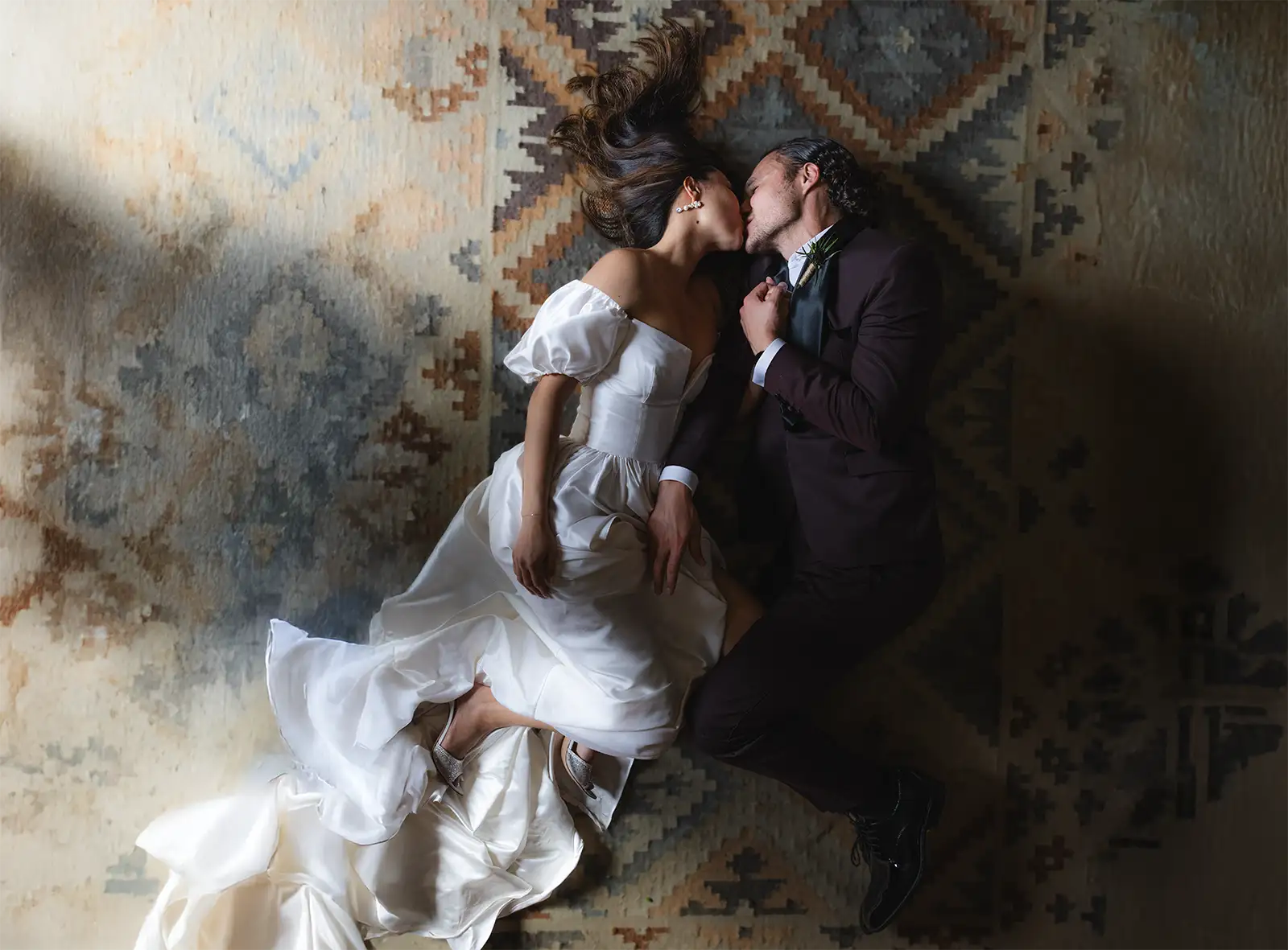 From overhead, a couple lie head to head kissing on a patterned rug, skirt fanned out, San Canzian.