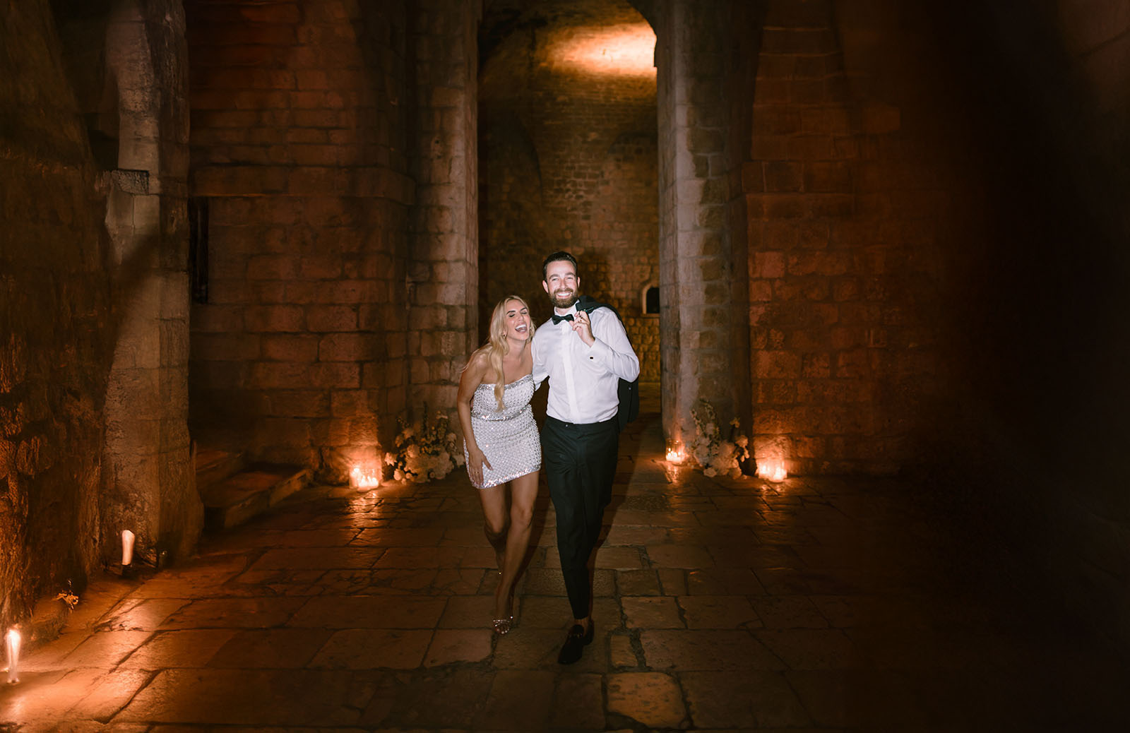 A couple walk through a candlelit stone hall of massive piers and arched vaulting, Fort Lovrijenac, Dubrovnik.