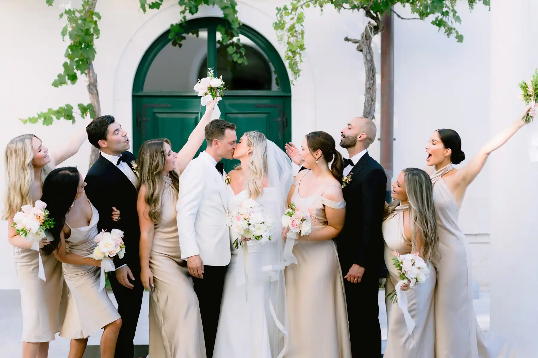 A couple kiss surrounded by guests lifting bouquets, a white wall behind them, Brown Beach House, Trogir.