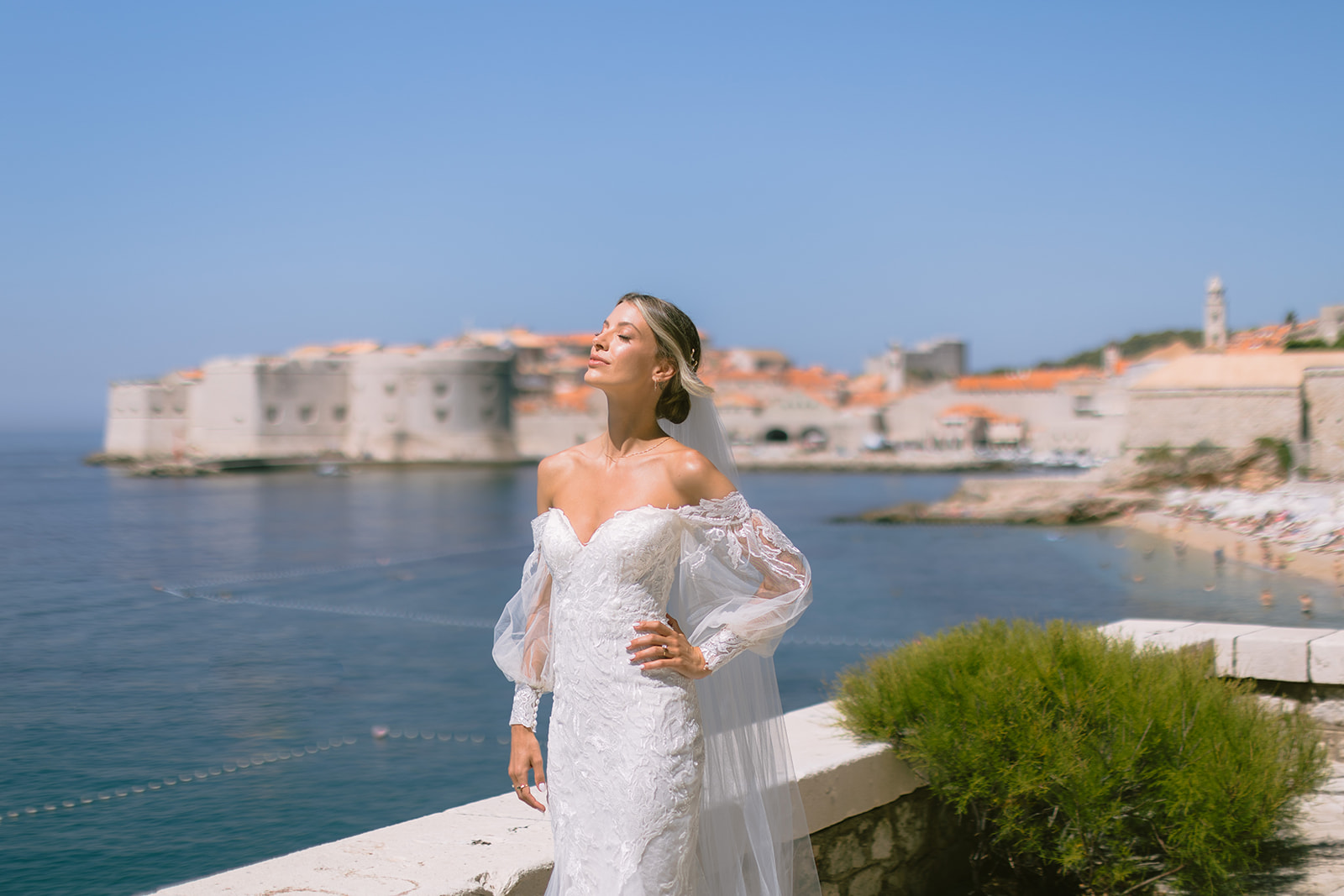 Bride alone on a sea-wall terrace, eyes closed, with Dubrovnik's Old Town and the public beach across the water behind her