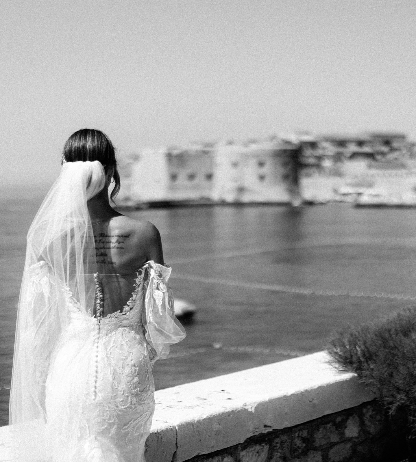 Black and white photo of a bride's back as she looks toward Dubrovnik's Old Town across the water