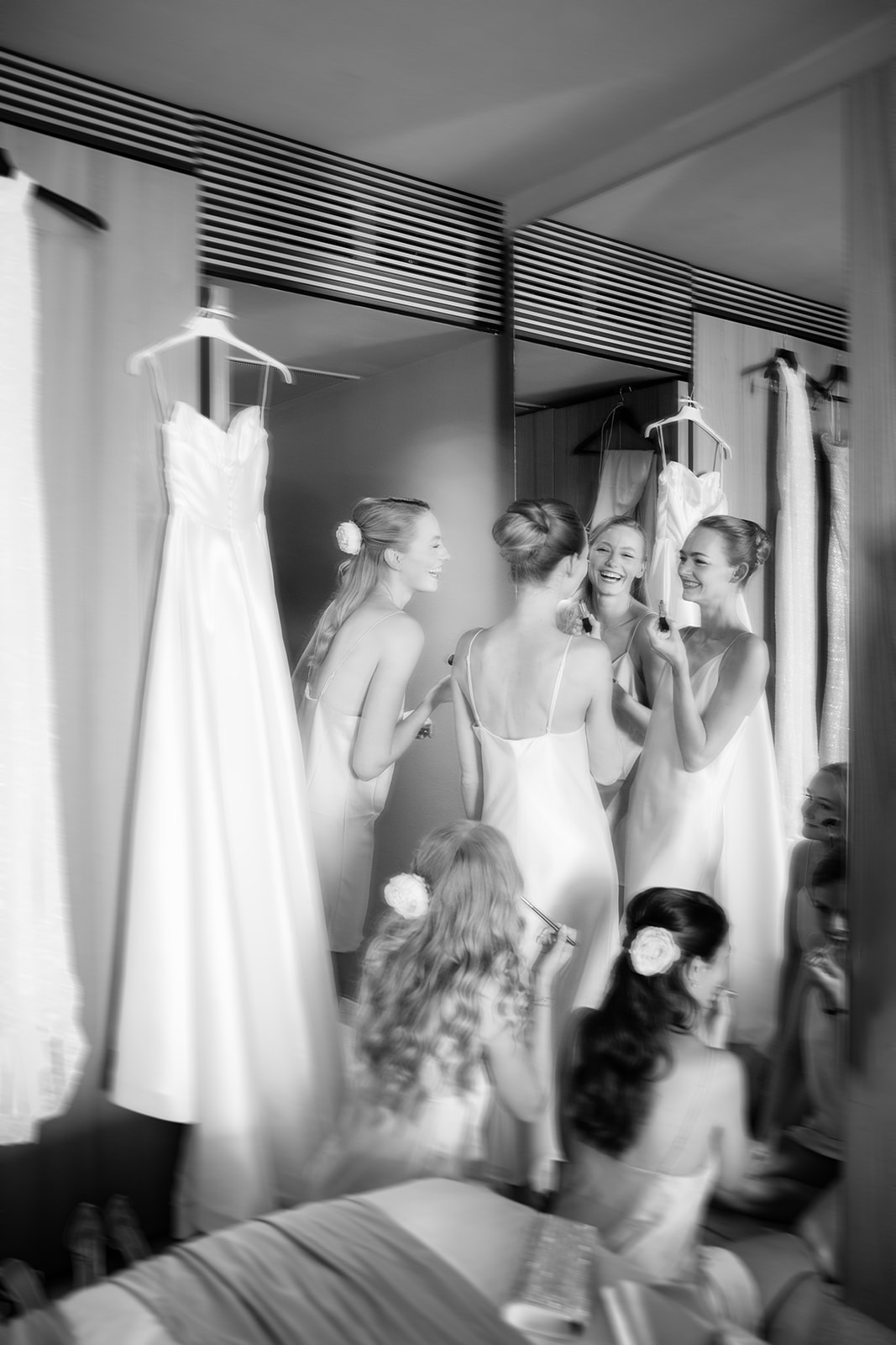 Bridesmaids in slip dresses laughing together while getting ready, with the wedding dress hanging nearby