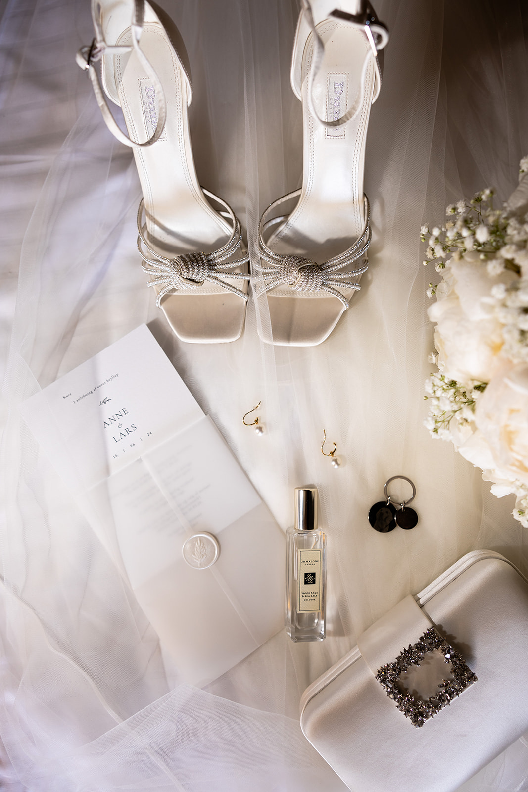 Flatlay of wedding-day details: shoes, invitation, jewelry, perfume and a bouquet corner on tulle fabric