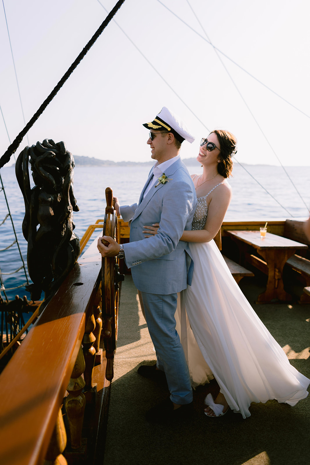 Bride and groom stand smiling at the bow of a sailing boat, the groom wearing a captain's hat