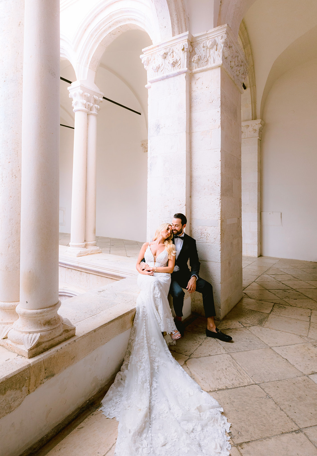 Bride and groom sit close together against a carved stone column inside a vaulted cloister