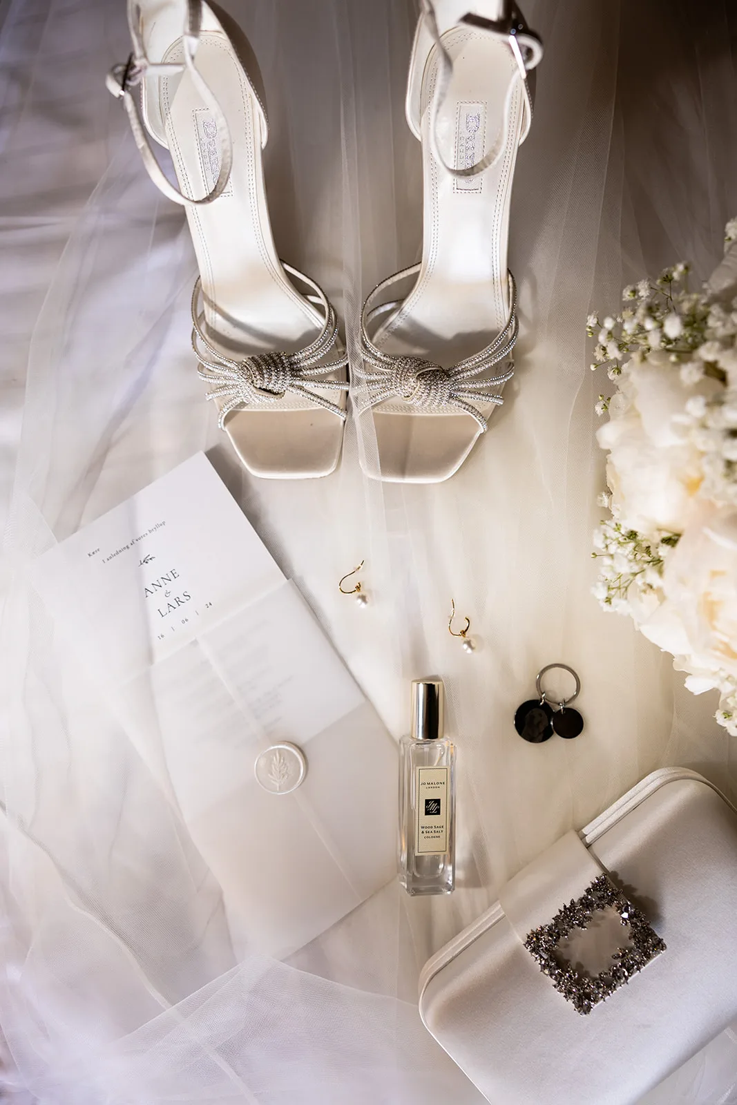 Wedding-day accessories including strappy rhinestone heels, a wax-sealed stationery card, perfume, pearl earrings and an embellished clutch arranged on a tulle veil beside a white peony bouquet corner.