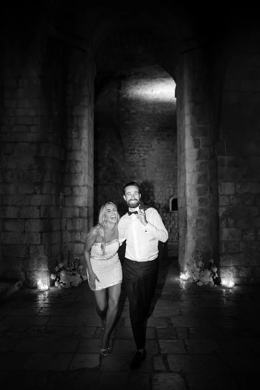Bride and groom walk arm in arm through a candlelit vaulted stone corridor at night, pillar candles and white floral clusters lining the columns.