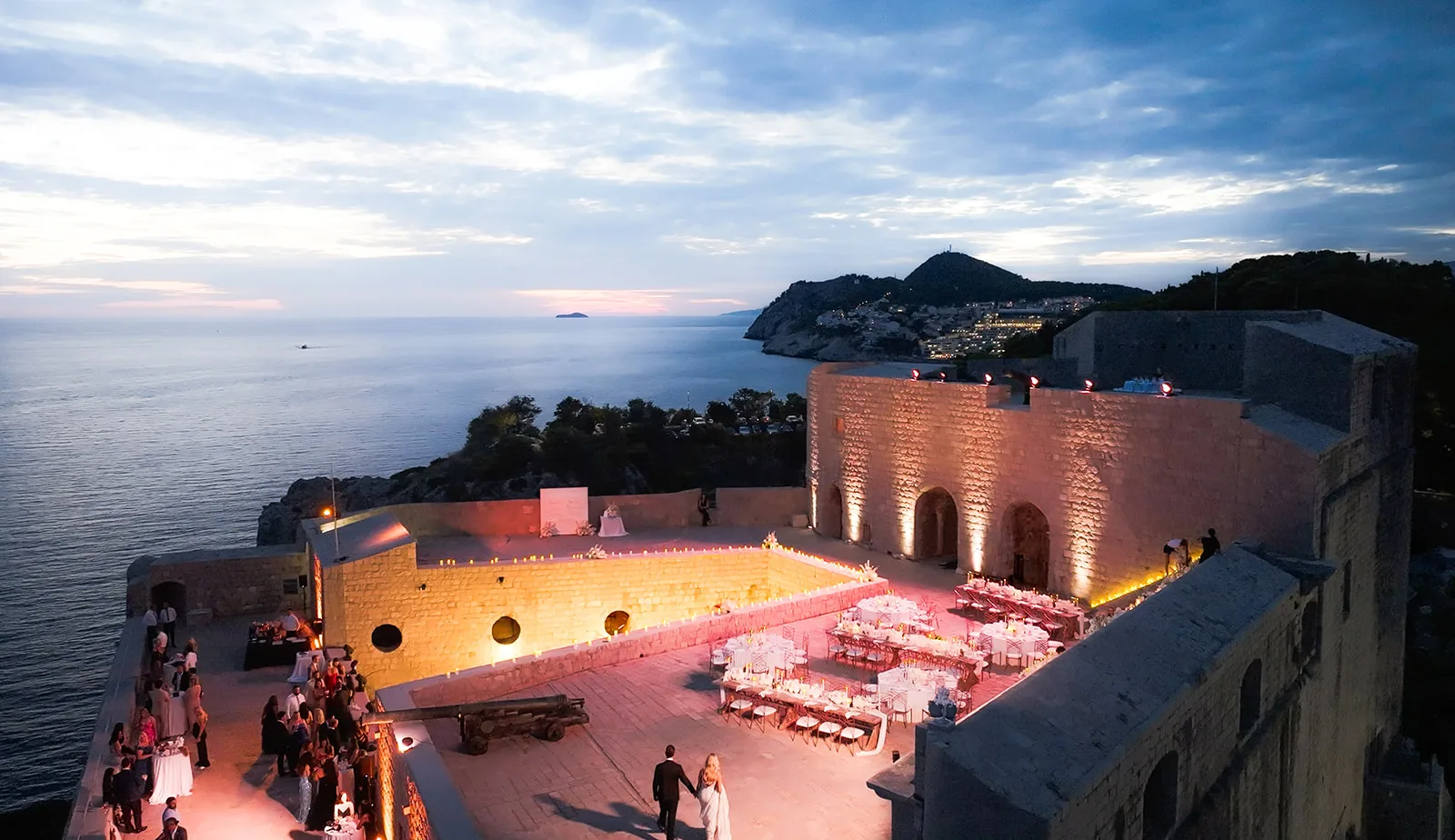 The couple stand alone on the upper terrace of a clifftop fort at dusk, looking down at their dinner table set in white linen, with guests still at cocktails on the level below and the Adriatic on three sides.