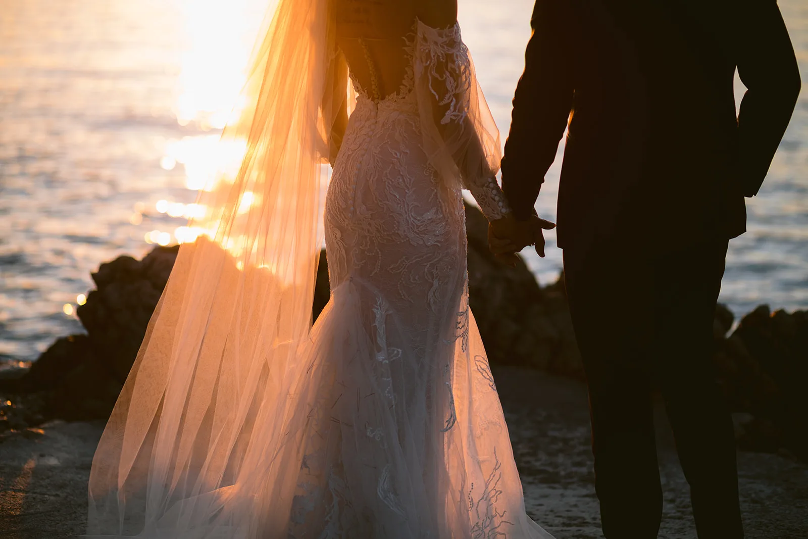 Backlit detail of the bride's lace gown and sheer veil sweeping over shoreline rocks, her hand linked with the groom's silhouette beside her.