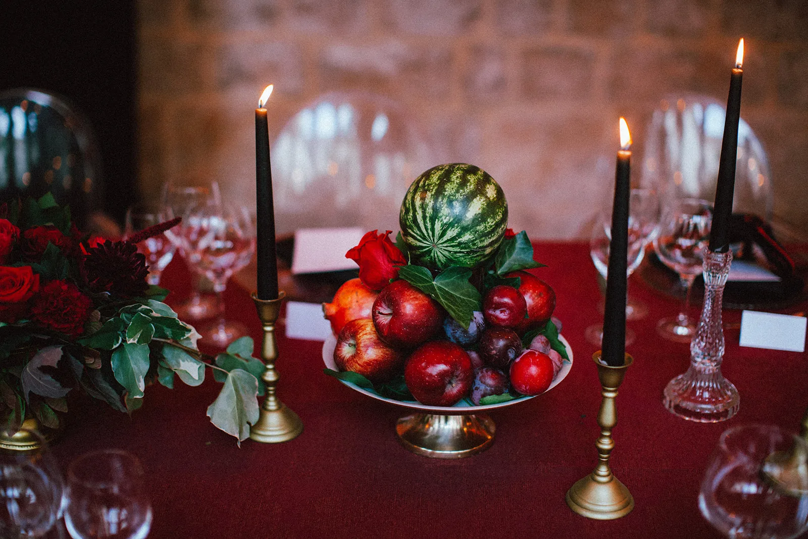 A candlelit still life of watermelon, apples, pomegranate and plums with a single rose on a red-draped table.