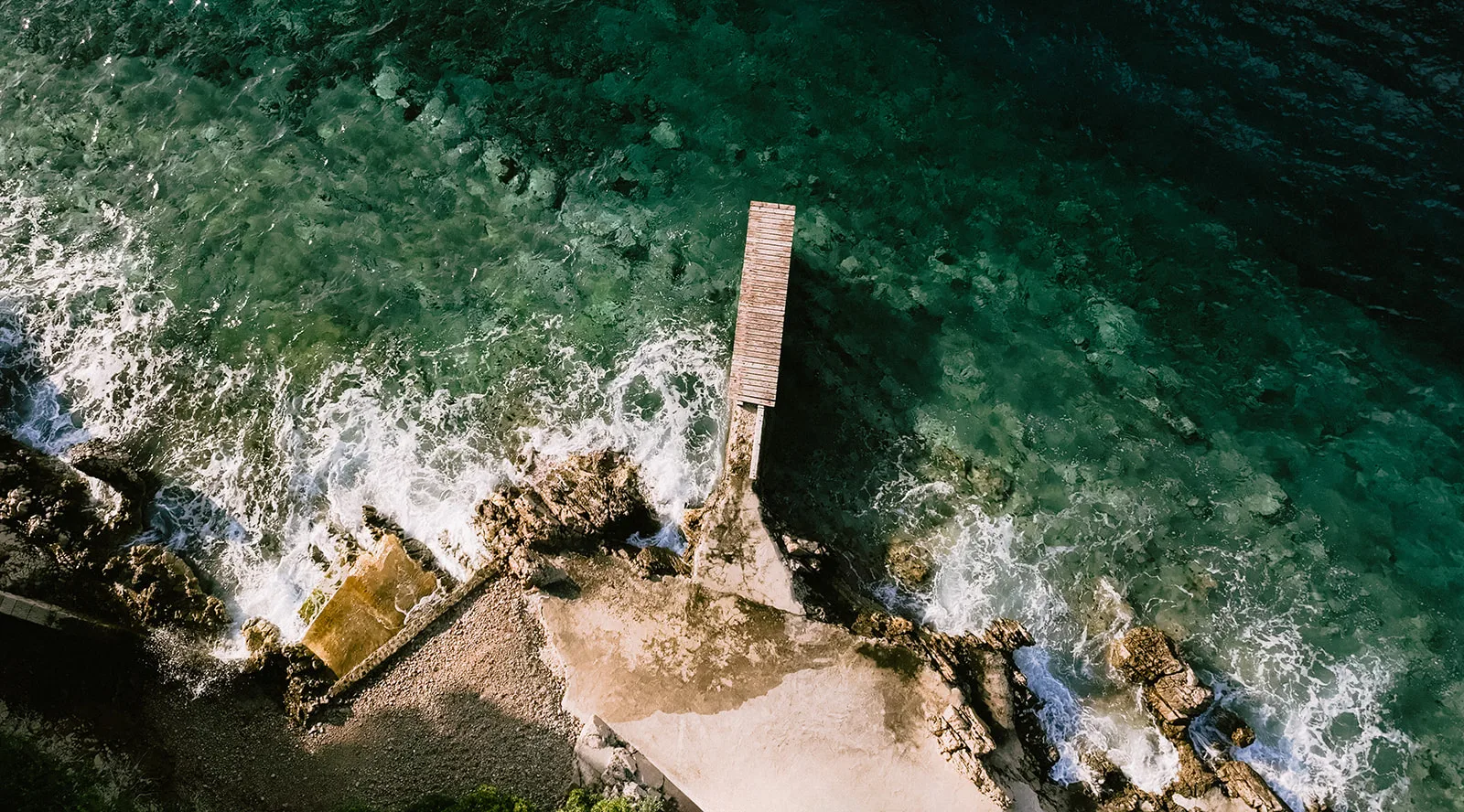 Aerial view of a wooden jetty reaching into turquoise sea at a rocky Adriatic shoreline, waves breaking white against the rocks below.