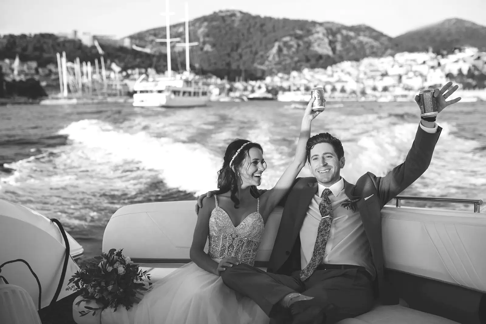 Bride and groom raise cans in celebration on speedboat stern as Hvar harbour town recedes behind in black and white