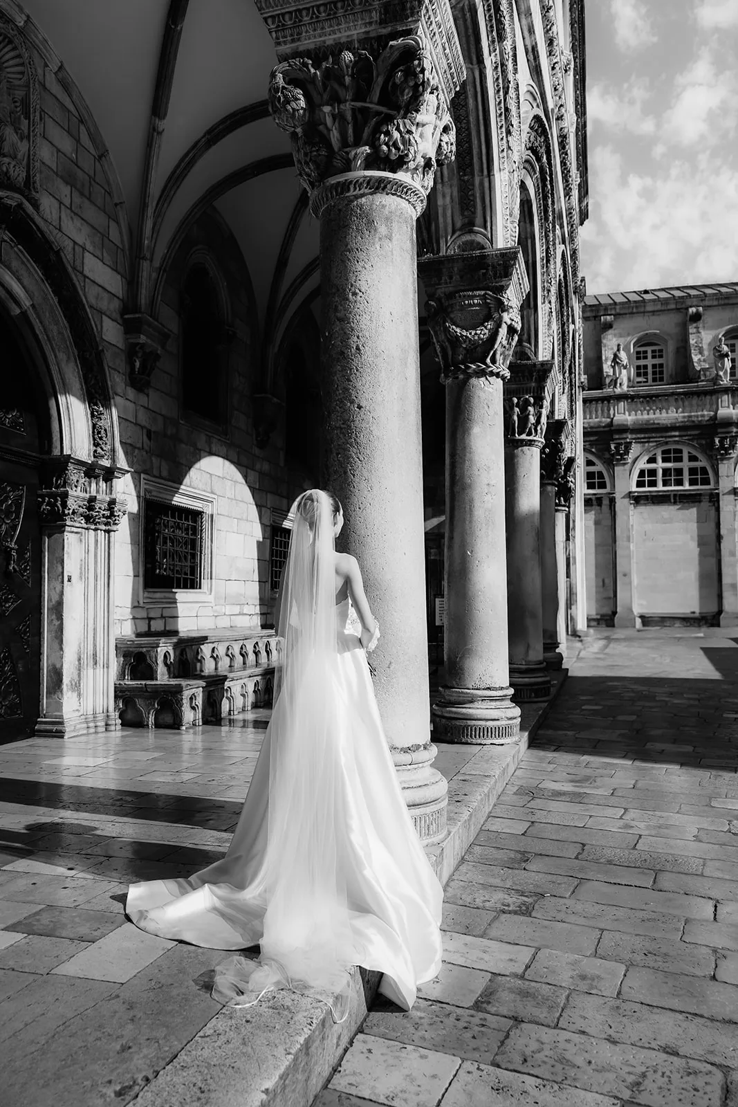 A bride is seen from behind walking through an arcaded stone loggia, her veil and dress train trailing over the paving, ornately carved column capitals framing her against a sunlit courtyard beyond, in black and white.