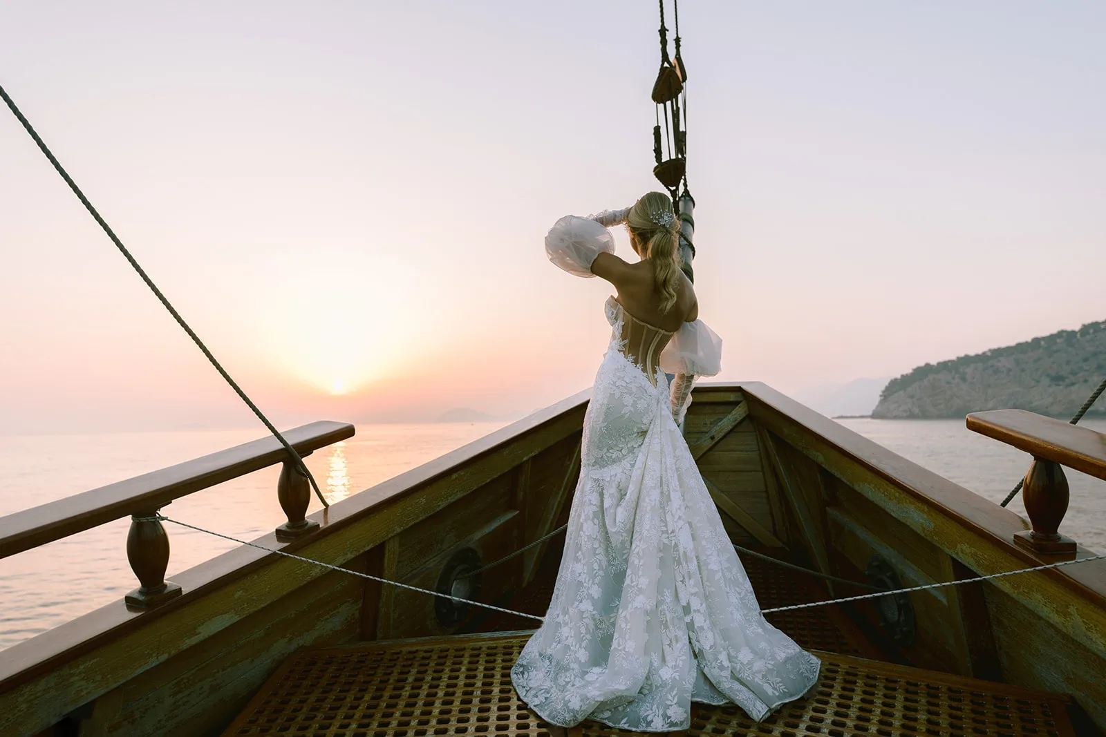 Bride stands alone at the bow of a wooden sailboat, back to camera, lace gown train pooling on the deck as she raises a hand to her hair, looking out at the setting sun.