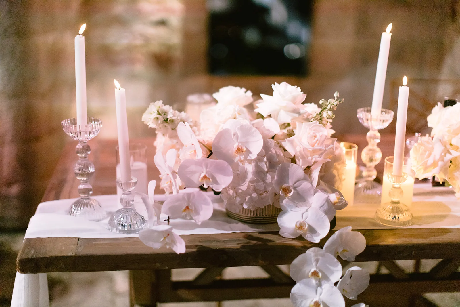 A row of crystal candlesticks and votive glasses beside white roses on the reception table.