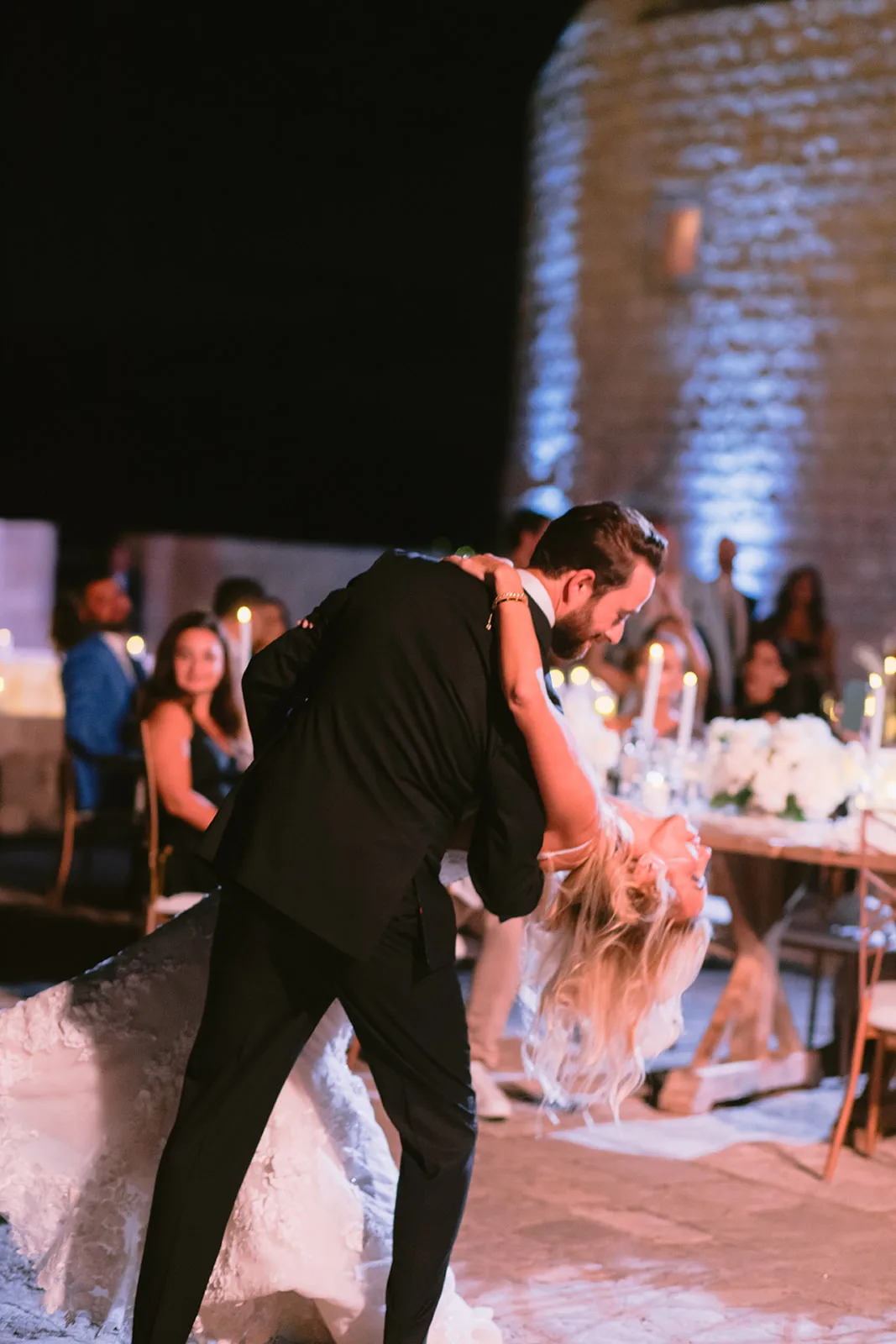 Groom dips the bride back in a dance move at night, her hair swinging loose, on a terrace in candlelight.