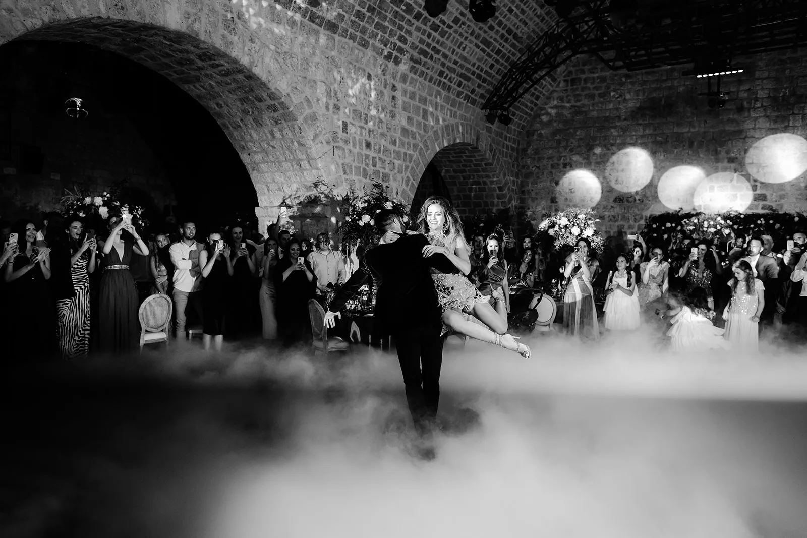 Groom lifts the bride off the ground mid-dance as guests crowd close around them.