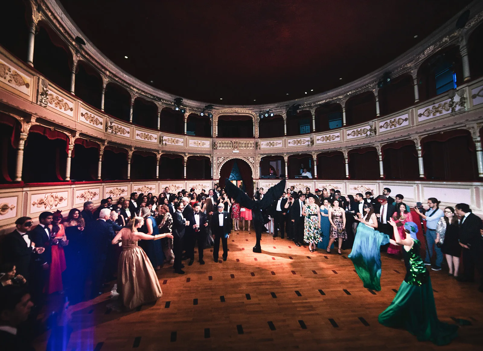 A costumed performer in large black wings stands at the centre of a grand theatre floor.