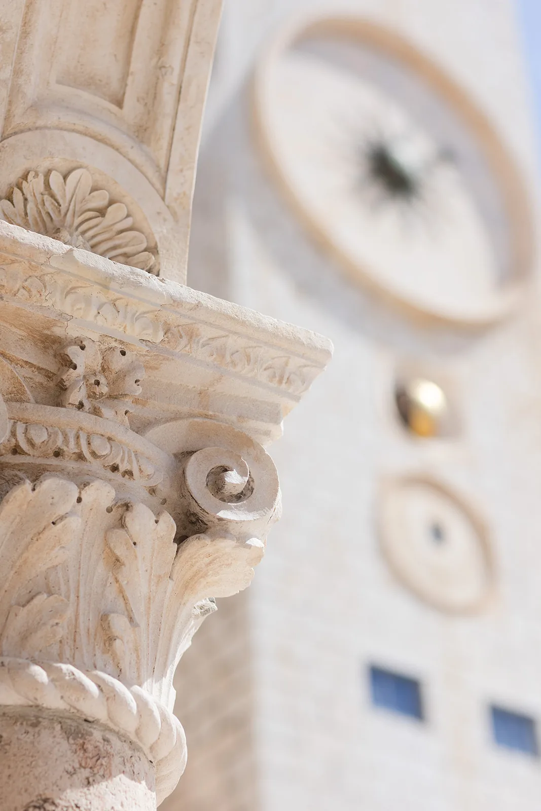 Extreme close-up of a carved acanthus-leaf column capital, sharp against a blurred bell tower beyond.