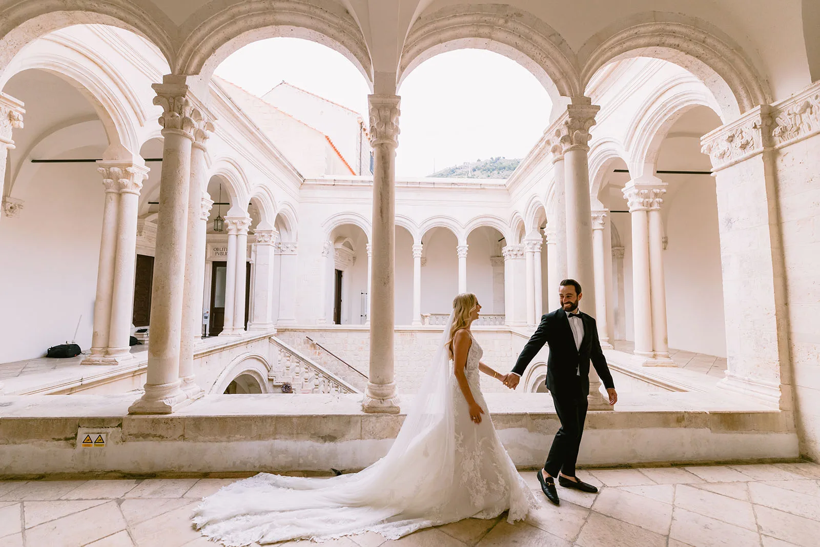 An empty grand stone atrium with a double staircase and an arcaded loggia of carved Renaissance columns, no one in it.