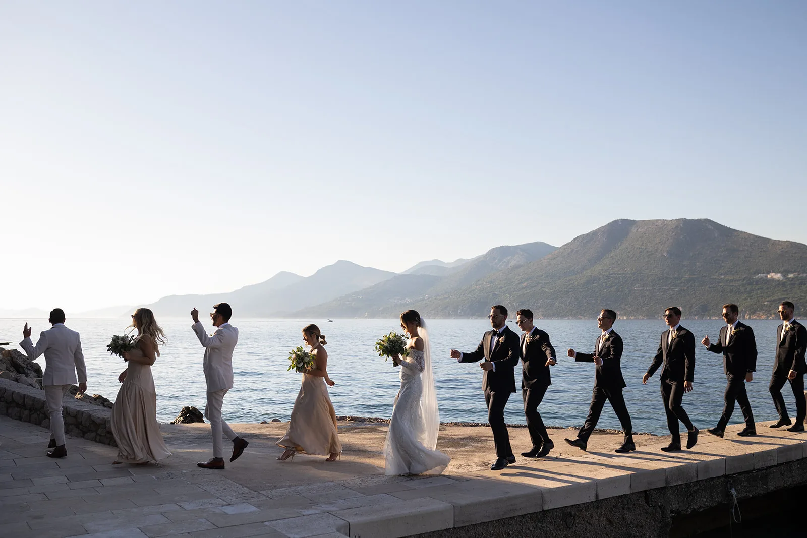 The wedding party walks single file along a stone breakwater above the sea, the bride and her bouquet at the centre, layered coastal mountains behind them.