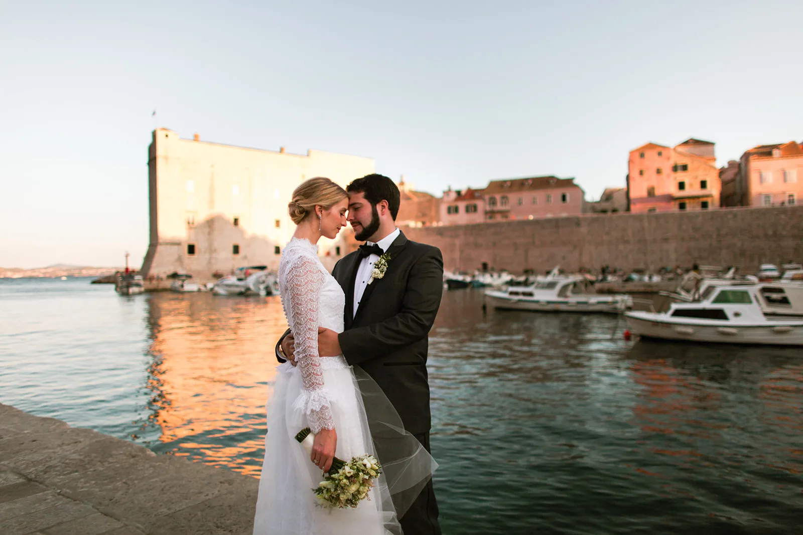 Bride and groom stand forehead to forehead on the Old Town harbour quay at sunset, the fort tower glowing warm behind the moored boats.