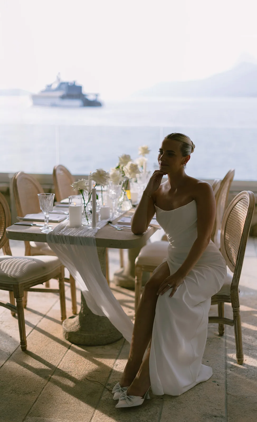 A bride sits at a candlelit dinner table on a seaside terrace, chin resting on her hand.