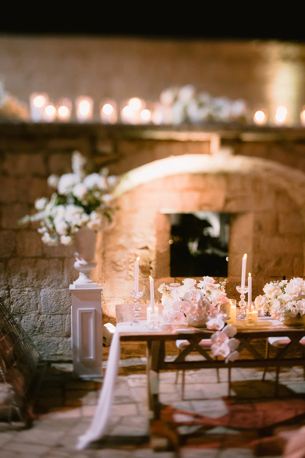 A candlelit reception table is set beneath a stone wall terrace, a white urn of flowers nearby.